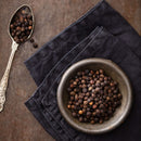 A metal bowl brimming with Tellicherry Peppercorns rests atop a dark cloth napkin beside an ornate spoon filled with more peppercorns, all arranged on a rustic brown surface—an ideal setup for refilling your The Spice Lab Premium Black Pepper Grinder by The Spice Lab.