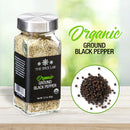 A glass jar labeled The Spice Lab Organic Ground Black Pepper, crafted from premium organic peppercorns by The Spice Lab, sits on a table. Next to it is a plate filled with whole black peppercorns, ideal for seasoning blends. On a light background, the text reads The Spice Lab Organic Ground Black Pepper.