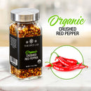 A jar of The Spice Lab Organic Crushed Red Pepper Flakes, an ideal pizza seasoning from The Spice Lab, is placed beside three fresh red chili peppers on a marble surface, with the product name shown in green and black text.