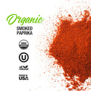 On the right, vibrant orange-red The Spice Lab Organic Smoked Paprika powder is scattered. On the left, bold text reads “The Spice Lab Organic Smoked Paprika” with USDA Organic, Kosher, All Natural, and Packed in USA icons displayed below. Experience its rich smoky-sweet flavor—ideal for BBQ rubs—all set against a crisp white background.