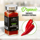 A glass jar of The Spice Lab Organic Smoked Paprika sits on a marble countertop, its label clearly visible. Beside it is a circle featuring two red chili peppers. Text reads: The Spice Lab Organic Smoked Paprika—prized for its rich smoky-sweet flavor, perfect for enhancing BBQ rubs and savory dishes.