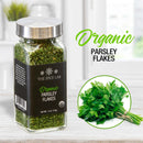 A glass jar labeled The Spice Lab Organic Parsley Flakes sits on a counter, displaying the vibrant green color of these premium dried herbs from The Spice Lab. Next to it is a photo of fresh parsley, emphasizing the natural quality and freshness of these perfect garnishing flakes. Text reads The Spice Lab Organic Parsley Flakes. The background features a bright kitchen scene with a window.