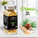 A glass jar labeled The Spice Lab Organic Pine Nuts rests on a counter. Nearby are pine needles, a pine cone, and pine nuts in a wooden scoop. Ideal for topping salads or as a baking ingredient, The Spice Lab’s organic pine nuts enhance your favorite dishes with pure, natural flavor. The text “The Spice Lab Organic Pine Nuts” appears in green and black lettering.