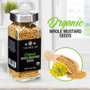 A glass jar of The Spice Lab Organic Whole Mustard Seeds is placed beside a wooden scoop filled with the same organic seeds and a small yellow mustard flower, perfect for pickling or brining, arranged on a light surface.