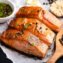 Three cooked salmon fillets garnished with fresh herbs and The Spice Lab Kings Blend Peppercorn Grinder Refill, served on parchment paper alongside a bowl of sauce, a wooden spoon, a halved garlic bulb, and a lemon slice in the background.