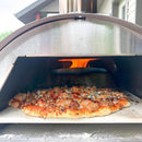 A pizza with sausage and sauce toppings bakes inside the WPPO Lil Luigi Portable Oven by WPPO LLC Direct, with an open flame visible in the background.