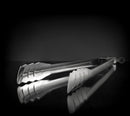 A pair of Napoleon Grills Stainless Steel Easy Locking Tongs (55011) rests on a glossy black surface, their reflection visible below, set against a dark, softly lit background.