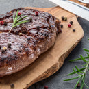 A grilled steak garnished with a sprig of rosemary and freshly ground pepper from The Spice Lab Premium Kings Peppercorn Grinder sits on a wooden cutting board, accompanied by a knife and decorative rosemary sprigs.