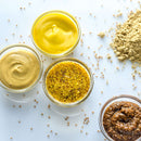 Four types of mustard in glass jars—including yellow, Dijon, whole grain, and spicy brown—are arranged on a white surface with The Spice Lab Organic Whole Mustard Seeds scattered nearby, perfect for pickling or brining your favorite foods.