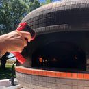 A hand holds the WPPO High Temp Infrared Thermometer for Wood-Fired Ovens by WPPO LLC Direct, checking the temperature of a wood-fired pizza oven with flames visible inside and trees in the background.