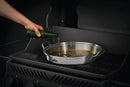 A hand pours liquid from a green bottle into a Napoleon PRO Chicken Infusion Roaster (56067) by Napoleon Grills, set on a grill.
