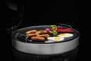 Sausages, eggs, onions, chili peppers, and herbed butter atop veggies sizzle on the Napoleon Cast Iron Reversible Griddle (56083) for 22" charcoal grills by Napoleon Grills, shown against a black background.