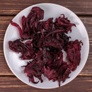 A white plate filled with dried, dark red The Spice Lab Whole Hibiscus Flowers—a sought-after botanical ingredient from The Spice Lab—sits on a wooden surface.