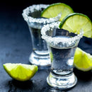 Two tequila shots with salted rims and lime wedges rest on a dark surface. The Spice Lab Traditional Margarita Salt Cocktail Rimmer by The Spice Lab has perfectly coated the glasses in margarita rim salt, while two lime wedges are placed nearby.