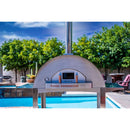 A WPPO Karma 55 304 Stainless Steel Commercial Wood-Fired Oven by WPPO LLC Direct is set up beside a swimming pool, surrounded by green trees and patio furniture beneath a blue sky.