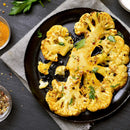 A roasted cauliflower steak garnished with herbs, spices, and a touch of The Spice Lab Organic Ground Mustard is served on a black plate, accompanied by spice bowls and parsley on a dark surface with a linen napkin.