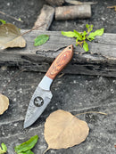 The Texan Knives 8" Forged Damascus Skinner Knife features a wire-cut blade and pakkawood & steel bolster handle, shown resting on a weathered log amid dried leaves and green foliage, alongside its leather sheath.