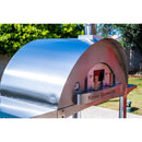 A WPPO Karma 55 304 Stainless Steel Commercial Wood-Fired Oven by WPPO LLC Direct is shown on a patio with trees and greenery in the background.