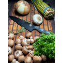 A cutting board featuring the Spaceman Knives Dark Matter Kiritsuke, surrounded by brown mushrooms, parsley, an onion, garlic, thyme, and a bundle of asparagus.