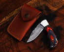 A Personalized Damascus Steel Pocket Knife by Texan Knives, featuring a folding blade and wooden handle, rests open beside its brown leather sheath on a carved wooden surface—a perfect custom Valentine’s Day gift for your husband.