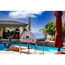 A WPPO LLC Direct Karma 55 304 Stainless Steel Commercial Wood-Fired Oven is positioned on a patio beside a blue swimming pool. The scene features a bright, sunny backyard with lush green trees, a covered patio area in the background, and a closed red umbrella on the right.