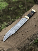 The Texan Knives Authentic Crocodile Dundee Knife features a 16” handmade Damascus steel blade and a buffalo horn or leather handle with brass guard, shown resting on a weathered log outdoors with blurred greenery in the background.