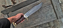A hand holds the Texan Knives Crocodile Dundee Outback Bowie Knife 2025, featuring a 16” handmade Damascus steel blade and leather/brass handle, above a weathered wooden chest. Includes sheath.