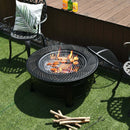 The DirectDeals Outdoor Fire Pit with BBQ Grill and High-temp Resistance Finish cooks food over flames on green grass, with chairs around it. Wooden sticks, grilling tools, and drinks are set nearby on a wooden deck.