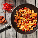 A frying pan filled with fried potatoes, sausage slices, and red bell pepper is perfectly seasoned with The Spice Lab Organic Spanish Sweet Paprika. Nearby, bowls hold extra red pepper strips and sausage slices, all arranged on a rustic wooden surface.