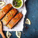 Three cooked salmon fillets garnished with herbs, pepper, and a sprinkle of The Spice Lab Organic Minced Garlic rest on parchment paper, accompanied by lemon slices and a small bowl of green herb sauce on a blue surface. Peppercorns and lemon slices are scattered nearby.