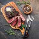A sliced steak with a pat of butter and rosemary is served on a wooden board, surrounded by The Spice Lab Kings Blend Peppercorn Grinder Refill from The Spice Lab, fresh herbs, a fork, and a knife on a dark stone surface.