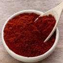 A white bowl filled with vibrant red The Spice Lab Organic Smoked Paprika, with a wooden spoon scooping up some of the spice, sits atop a light wooden surface. This flavorful seasoning by The Spice Lab is perfect for bringing a smoky-sweet taste to BBQ rubs and your favorite dishes.