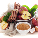 A bowl of red apples with green leaves, cinnamon sticks, brown sugar in a small bowl, a cracked egg, and a white cloth on a white background evoke the warm aromas of baking—perfect for crafting cozy treats with The Spice Lab Apple Pie Spice Blend from The Spice Lab.