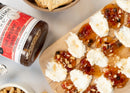A wooden board features goat cheese topped with Terrapin Ridge Farms Hot Pepper Bacon Jam 1 Gallon Jug, offering sweet heat and chopped nuts. Surrounding it are crackers, a jar of the jam, and a bowl of snack mix on a light surface.