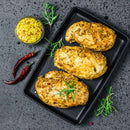 Three seasoned chicken breasts rest on a black rectangular plate, garnished with rosemary and accompanied by two dried red chilies. Beside them sits a small bowl of whole grain mustard made with The Spice Lab Organic Whole Mustard Seeds. All items are beautifully presented on a dark stone surface.