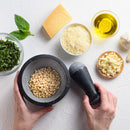 A hand holds a mortar and pestle containing The Spice Lab Organic Pine Nuts, surrounded by freshly chopped basil, Parmesan cheese, garlic cloves, olive oil, vibrant basil leaves, and a whole garlic bulb on a white surface—delivering perfect ingredients from The Spice Lab for homemade pesto sauces or as a delicious baking ingredient or salad topper.