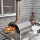A freshly baked pepperoni pizza sits on a metal peel, partially out of the WPPO Lil Luigi Portable Oven by WPPO LLC Direct, surrounded by snow on a patio next to a house with gray siding.
