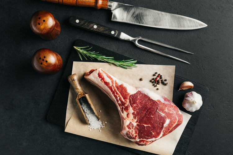 Raw ribeye steak on parchment paper with rosemary, garlic, peppercorns, salt, a meat fork, knife, and wooden salt and pepper shakers on a dark background.