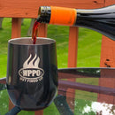 Red wine is poured from a Ríunite bottle into a black WPPO LLC Direct WPPO Hot and Cold Tumbler With Lid, featuring the GET FIRED UP logo. The stainless steel tumbler sits on a glass patio table outdoors, framed by a blurry green yard and an orange wooden deck railing in the background.