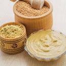 A bowl of creamy mustard, a wooden container of mustard seeds, and another with The Spice Lab Ground Yellow Mustard 1 lb—a favorite spice among foodservice professionals and perfect for adding tangy zest to BBQ seasoning—are arranged on a light wooden surface.