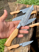 A hand holds a Texan Knives Sharp Kitchen Knife with a 13" Damascus steel blade and olivewood handle. More knives from the set are displayed outdoors on rustic wood, with grass and soil in the background. Leather sheaths are also shown.