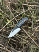 The Texan Knives Hunters Knife features a 9" D2 steel blade and an epoxy pine cone resin handle, displayed outdoors on dry branches and leaves, and comes with a leather sheath.
