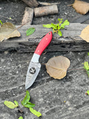 The Texan Knives FIXED BLADE POCKET KNIFE, a personalized Damascus steel hunting knife with a red handle, rests on dark stone among dry and green leaves and weathered wood.