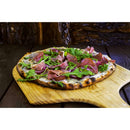 A wood-fired pizza topped with arugula and prosciutto rests on the WPPO 12" x 24" Wooden Pizza Peel (Launch Pad) from WPPO LLC Direct, set atop a rustic wooden table with a backdrop of stacked logs.