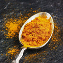 A silver spoon filled with bright orange The Spice Lab Turmeric Powder with Curcumin, from The Spice Lab, rests on a dark stone surface, with some of the vibrant powder scattered around—a scene that highlights the essential role of this turmeric powder in global cooking.