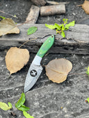 The Texan Knives Handmade Skinner Knife features an 8" Damascus steel blade and a pakkawood wire-cut handle, displayed on dark stone beside leaves, branches, and its rugged leather sheath.
