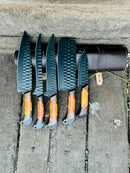 Five black Damascus steel chef knives with orange-accented wooden handles by Texan Knives are arranged on wood beside a leather case and sharpener—a perfect handmade gift for wife, husband, or Valentine’s Day.