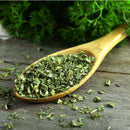 A wooden spoon filled with The Spice Lab Whole Leaf Cilantro rests on a rustic wooden surface, while fresh parsley leaves appear blurred in the background.