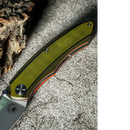 The Spaceman Knives Galatea Folding Pocket Knife by Spaceman Knives features a textured green handle with black and orange accents, visible screws, and a premium 14C28N steel blade. It rests on rough stone near bark in the image.