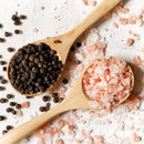 Two wooden spoons rest on a white surface: one is filled with The Spice Lab’s Premium Black Peppercorns, while the other holds crystals of The Spice Lab Himalayan Pink Salt. A sprinkle of salt and pepper surrounds the spoons, creating an inviting display for The Spice Lab Himalayan Pink Salt & Premium Black Pepper Grinder Set.
